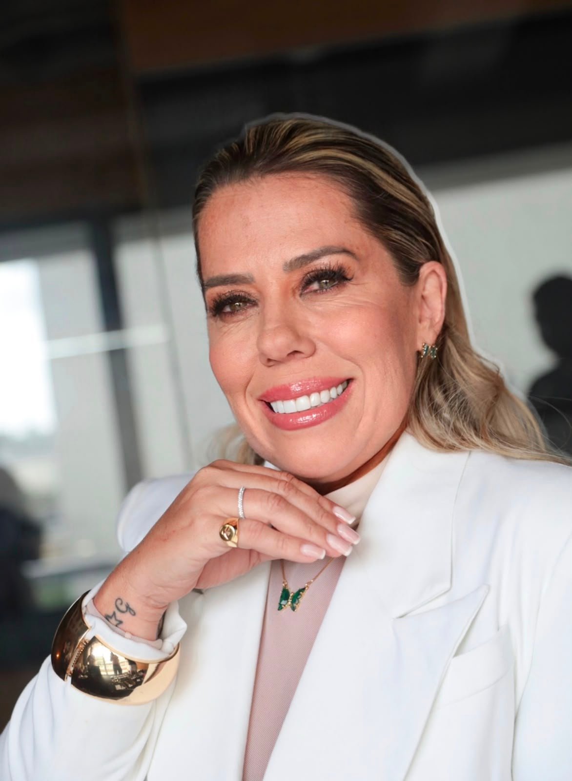 Daniela Amorim sorrindo, de blazer branco, com a mão apoiada no queixo
